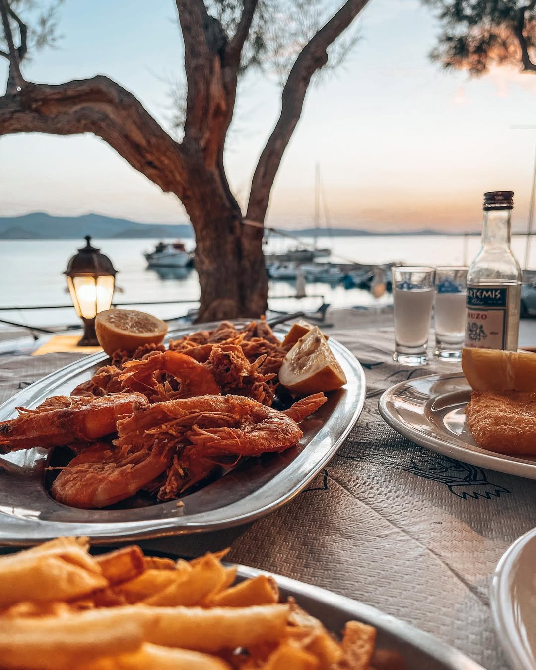 Large platter of golden-brown fried seafood including whole shrimp, calamari, and mixed seafood with lemon wedges, served at seaside Taverna with Artemis ouzo bottles