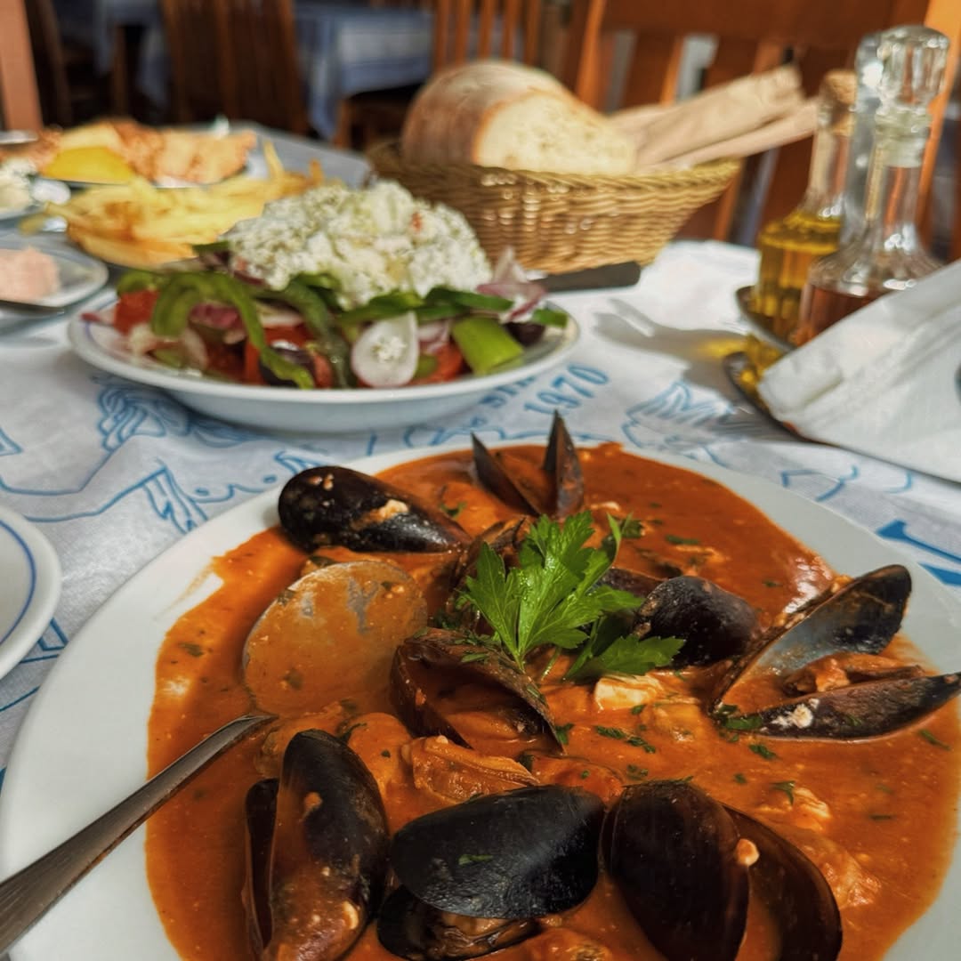 Fresh mussels in rich orange-red tomato sauce with parsley garnish, served on white plate at Gorgona Taverna with traditional Greek condiments and olive oil bottles